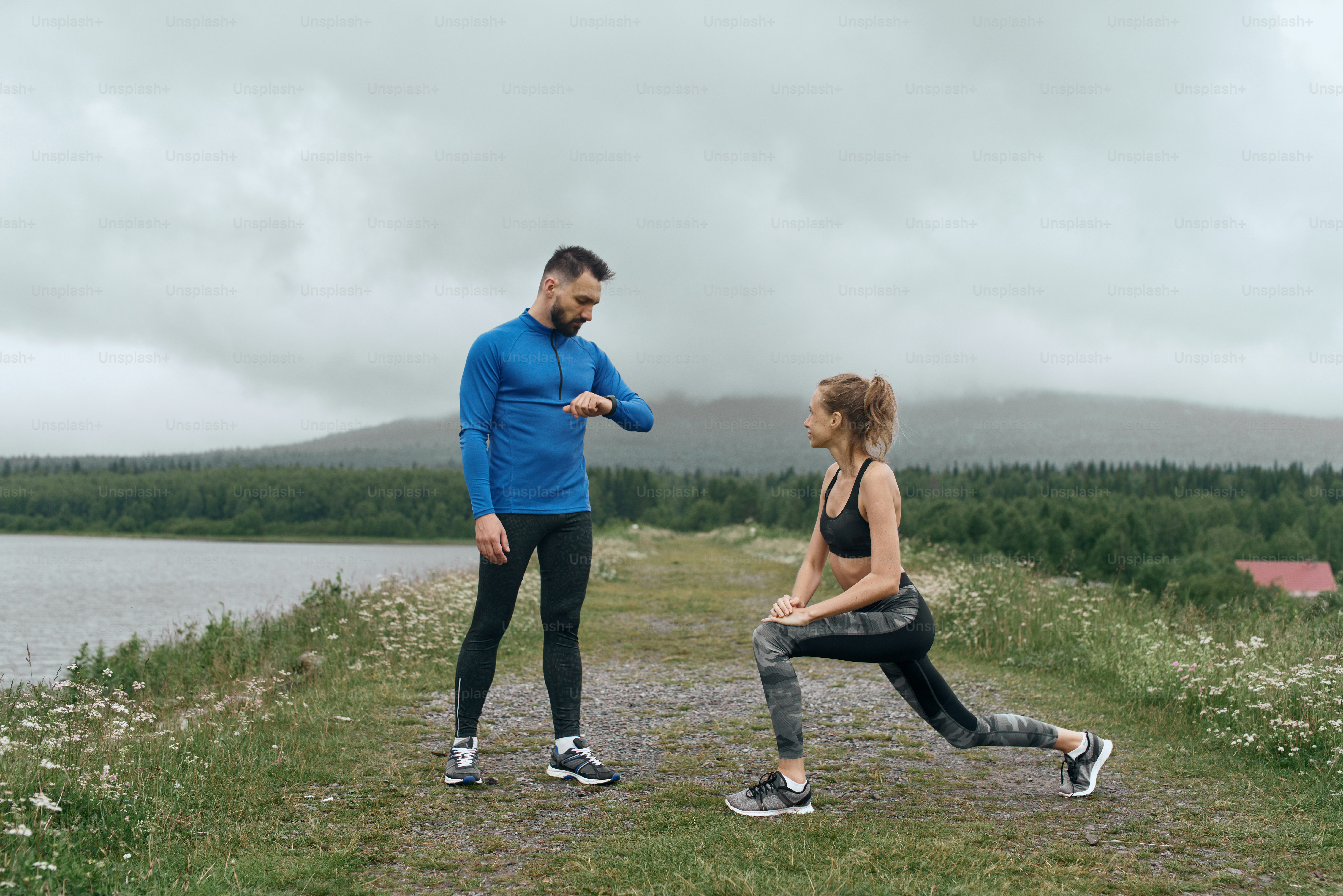 Two mid age man and woman exercising outdoor, warming up arm muscules, in summer, on gloomy day, standing at the road with scenic view, doing warm up stretching, wearing blue jacket, he has a beard