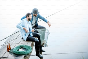 Two friends catching fish with fishing net and rod sitting on the wooden pier at the lake