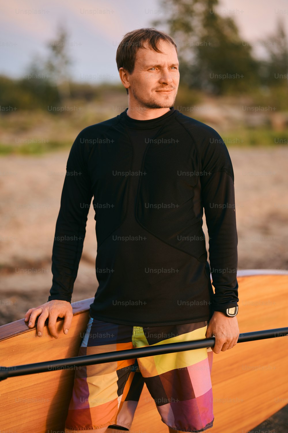 Portrait of strong man in wet shirt posing with sup surf board and paddle at the lake shore after active paddling on the water, looking away from camera