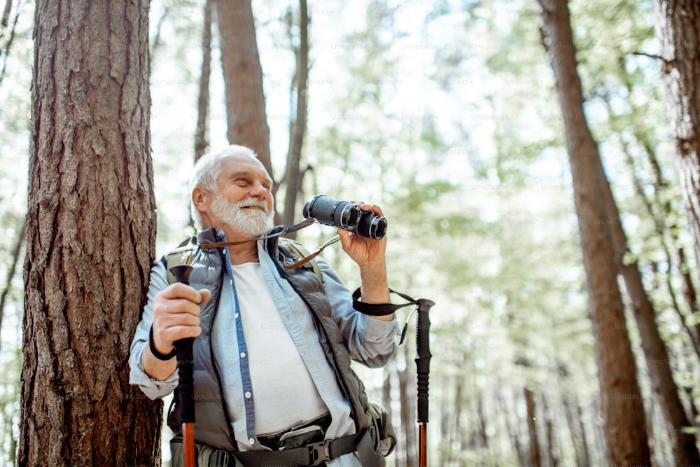Portrait of a senior man with binoculars and backpack resting near the tree while traveling in the forest