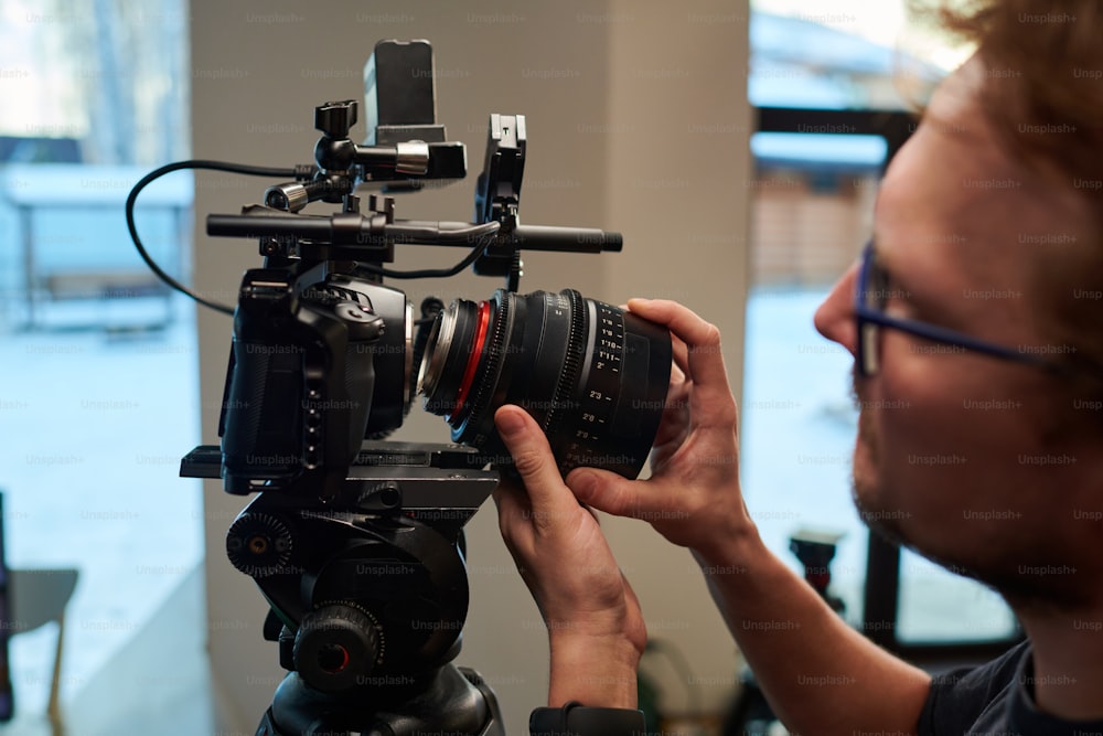 Hände eines jungen Kameramanns, der das Objektiv der Videokamera dreht, während er den Fokus einstellt und sich auf die Dreharbeiten für neue Werbung vorbereitet