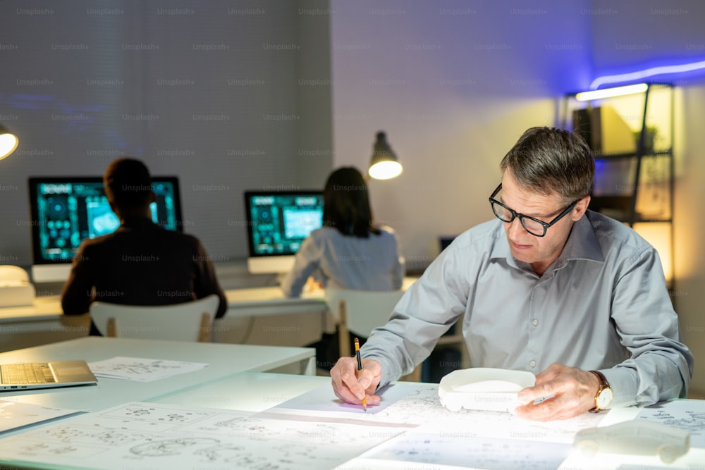 Concentrated mature automotive designer in eyeglasses sitting with car model at table and editing layouts in office