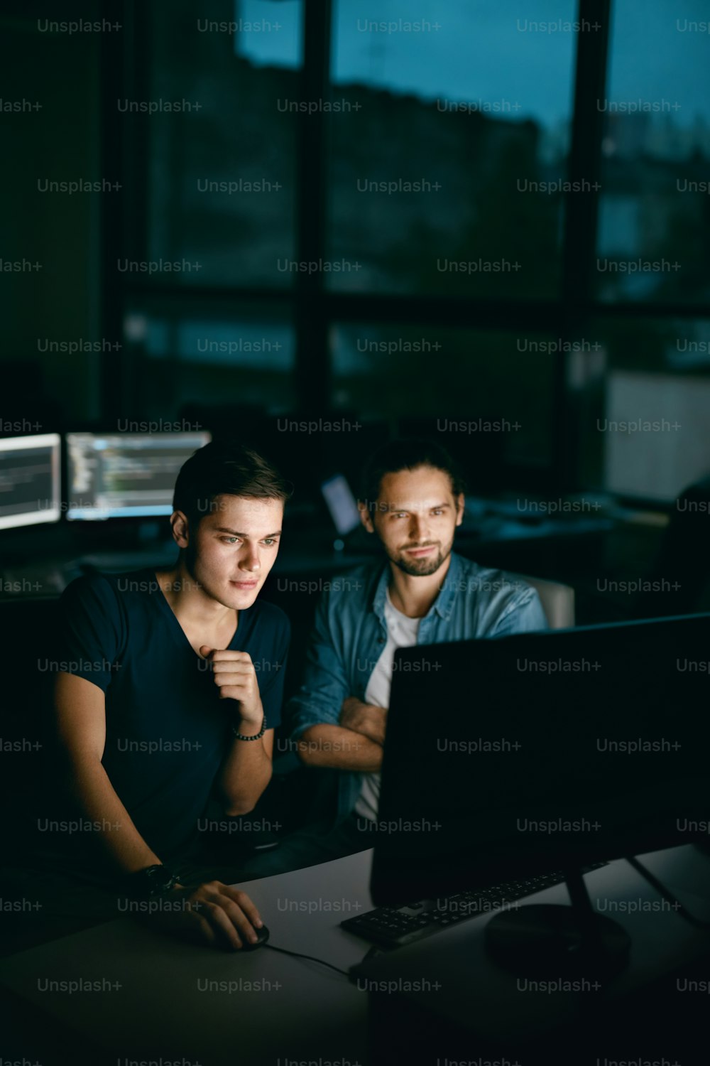프로그래머는 사무실에서 컴퓨터를 보고 일하고 있습니다. 캐주얼한 잘생긴 젊은이들은 저녁에 직장에 앉아 컴퓨터 작업을 하면서 코드를 입력합니다. 고품질 이미지.