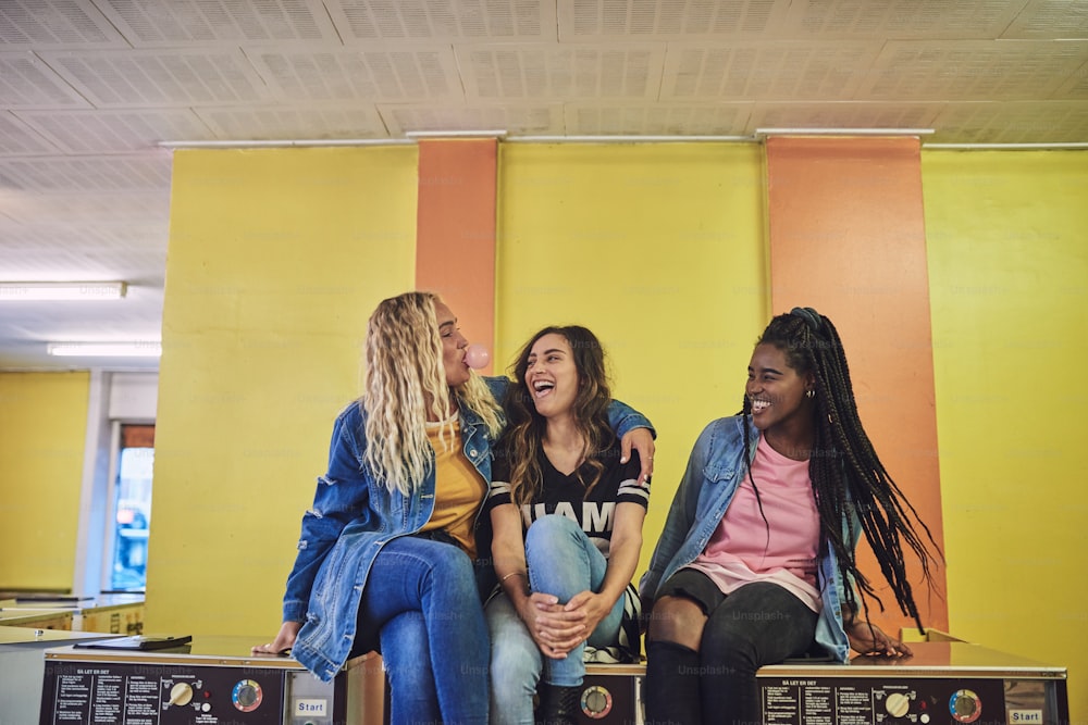 Three young girlfriends sitting together on washing machines in a laundromat laughing and blowing bubbles with chewing gum