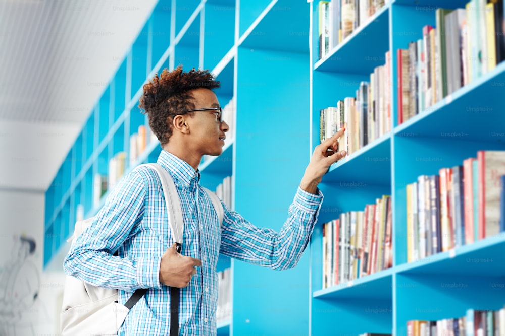 Clever teenage guy with backpack looking for necessary book or manual in college library