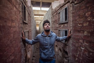 Portrait of focused motivated afro-american young handsome sportive man with earphones standing inside of the abandoned place in the middle of two walls and thinking while looking up.