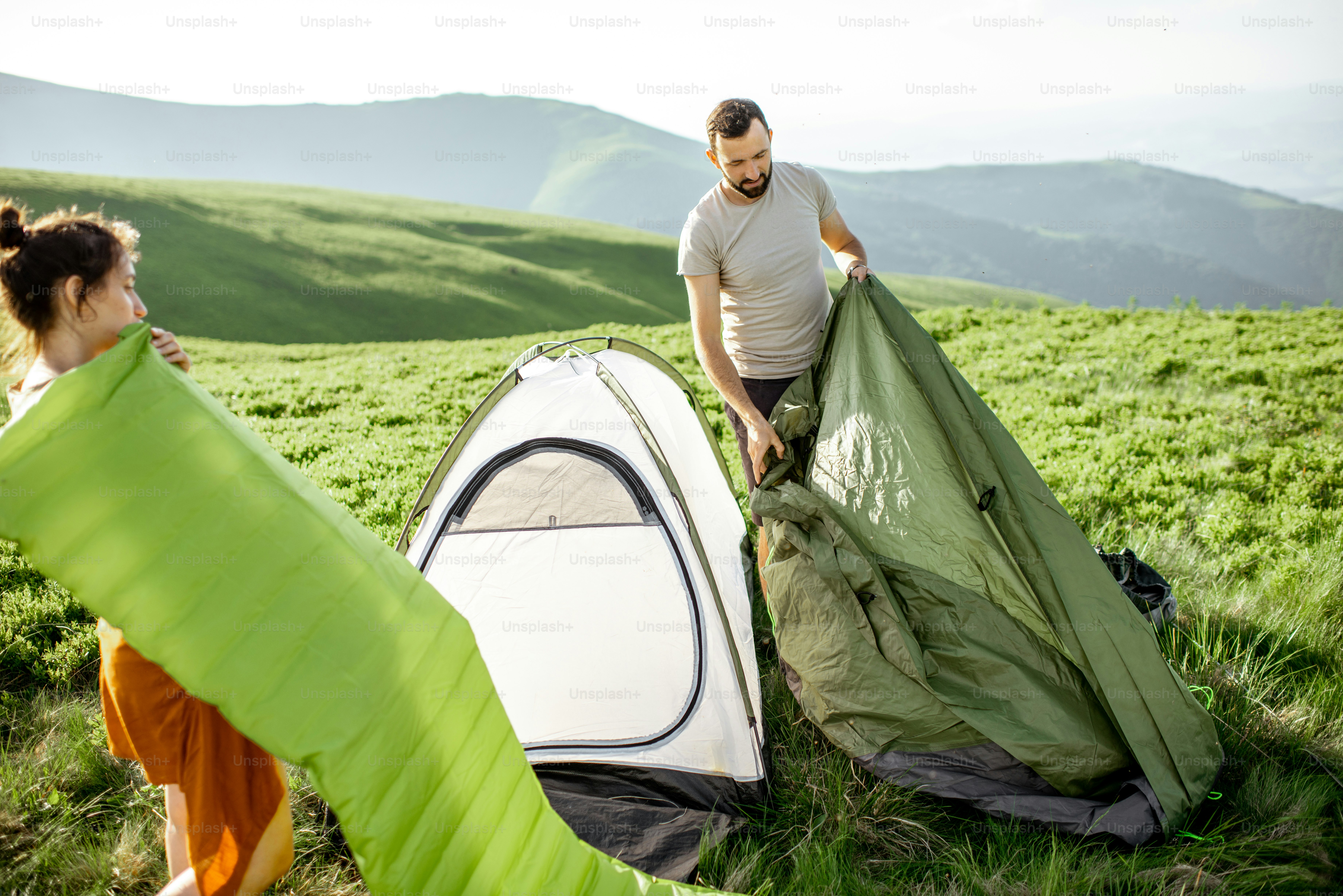Giovane coppia che monta la tenda e gonfia il materasso sul prato verde, viaggiando in alta montagna durante il tramonto