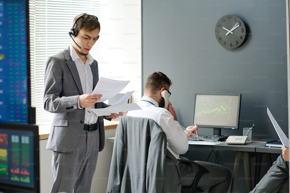 Two men in formalwear working in brokerage company having phone calls with their business partners and clients and looking through documents and stats