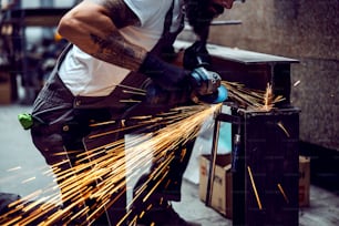 Nahaufnahme eines muskulösen tätowierten Arbeiters mit Handschuhen, der neben einem Metallbalken kniet und ihn mit einem Schleifer bearbeitet. Ein Arbeiter in der Werkstatt beim Schleifen von Metall