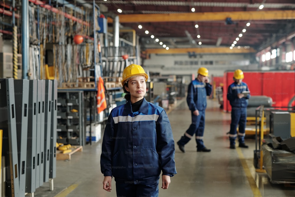 Serious confident young female factory foreman in hardhat walking along compressor plant shop and controlling work of employees