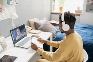 Joven estudiante africana con auriculares sentada frente a una computadora portátil durante la lección en línea y escuchando al maestro con hiyab