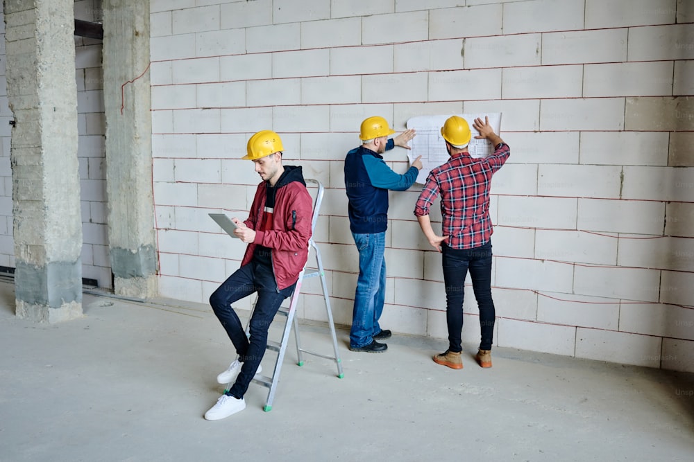 Junger Ingenieur mit Schutzhelm sitzt auf einer Trittleiter und scrollt mit seinen Kollegen durch die Beschreibung von Baumaterialien