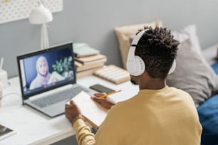 Young African male student in headphones looking at teacher on laptop screen explaining new subject during online lesson