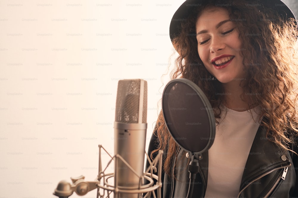 Ritratto di bella donna riccia in giacca di pelle e cappello che registra le voci in studio di musica su apparecchiature audio professionali