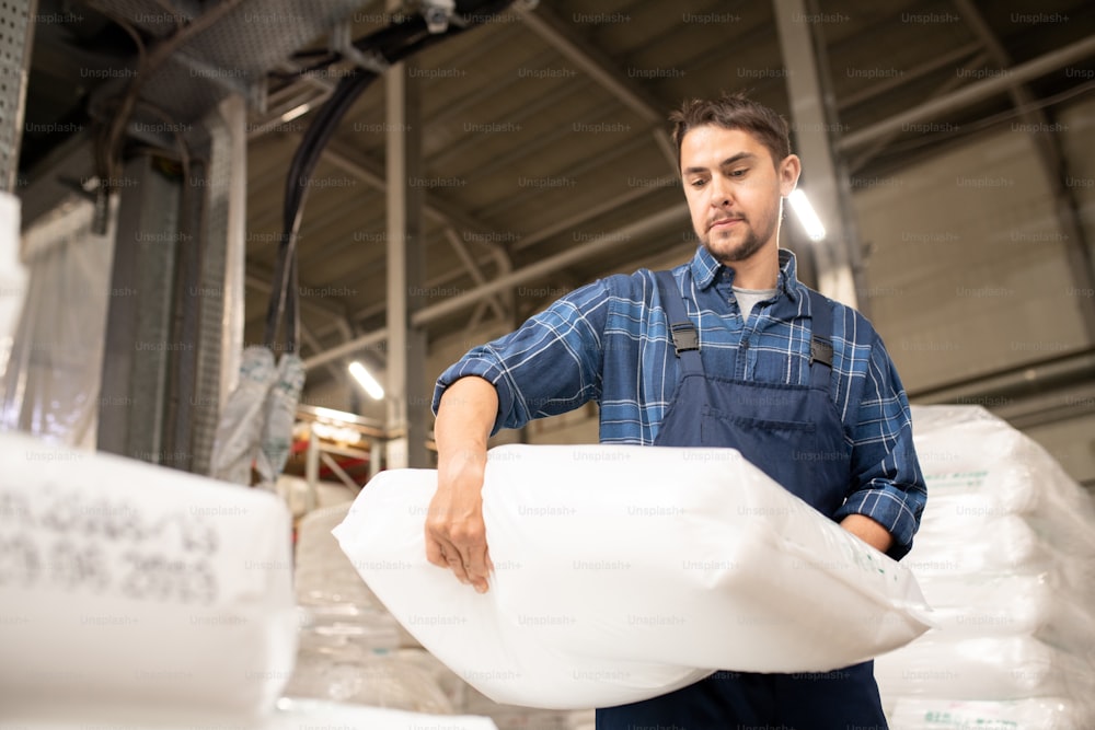 Hombre joven en ropa de trabajo que lleva un pesado saco blanco con gránulos de polímero durante el proceso de carga en una gran fábrica