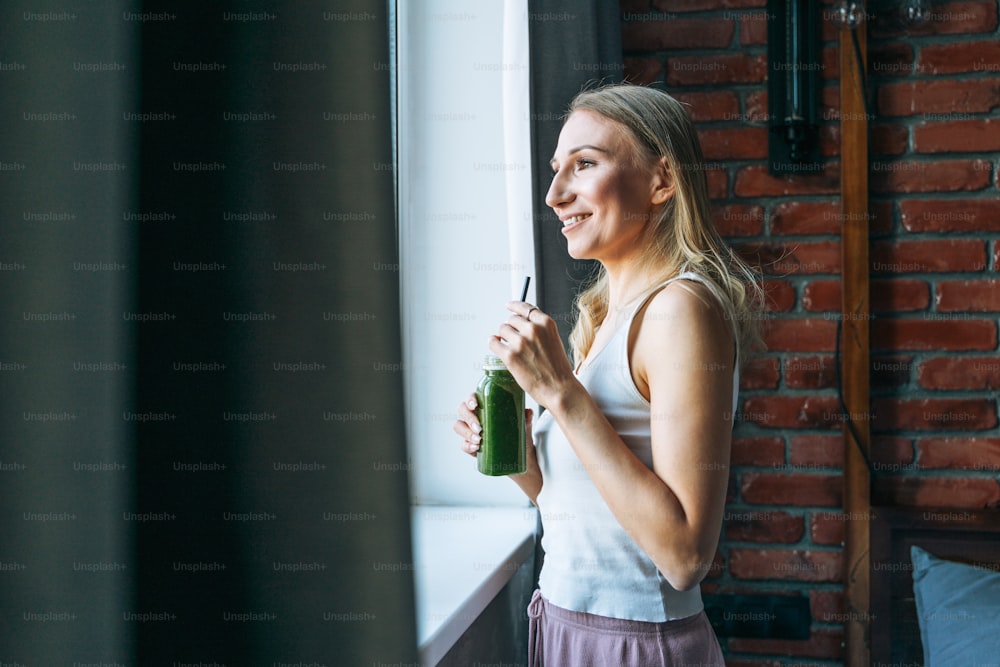 Young adult woman with blonde long hair in casual with green smoothie near window at home
