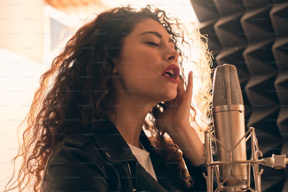 Portrait of beautiful curly woman in leather jacket and hat recording vocals in music studio on professional sound equipment