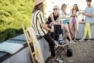 Stylish carefree persons talk and drink beer at roof top terrace at countryside house. Friends hanging out at picnic outdoors