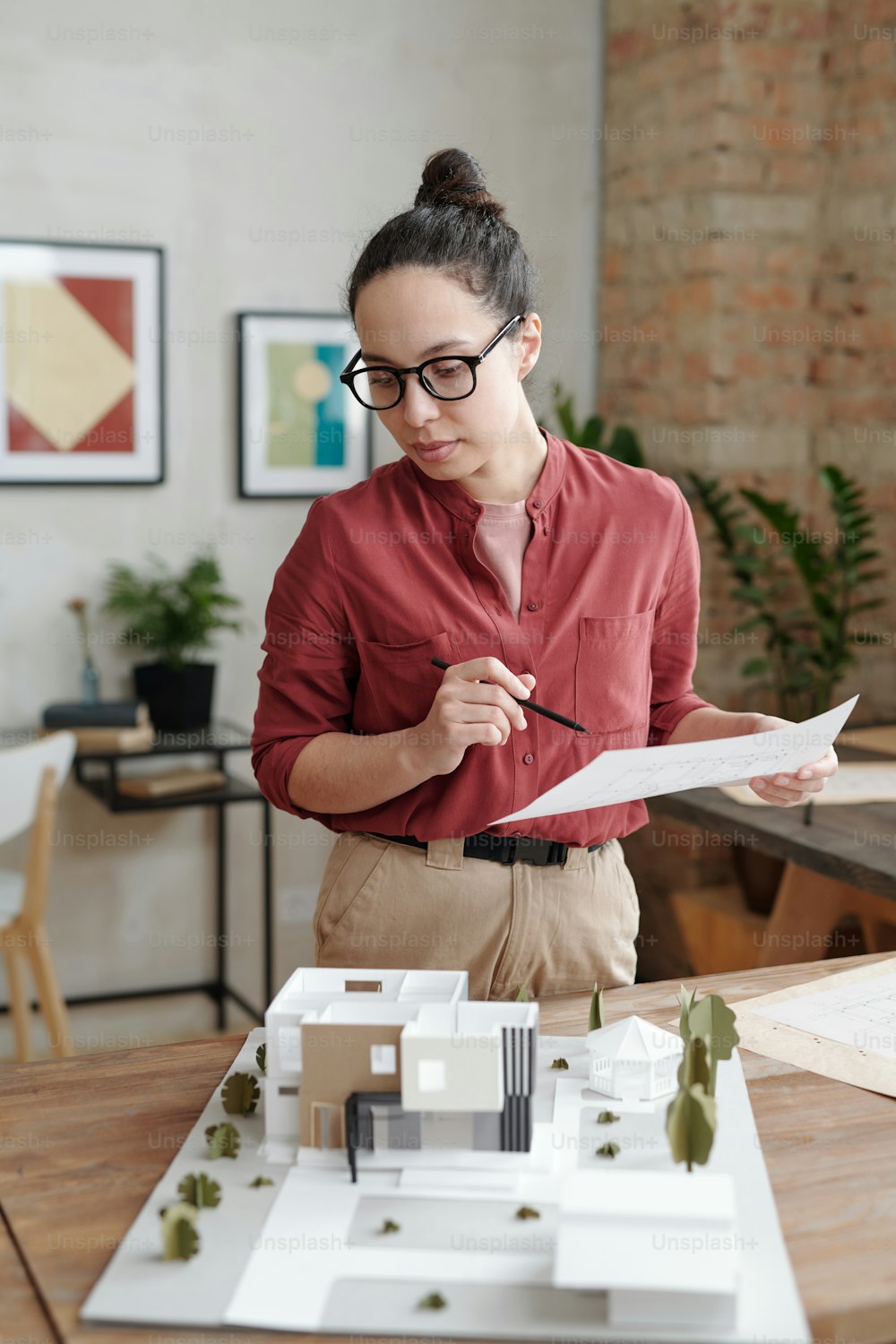 Giovane donna ispanica fiduciosa in occhiali da vista in piedi alla scrivania con maquette di edificio moderno e che analizza la planimetria della casa
