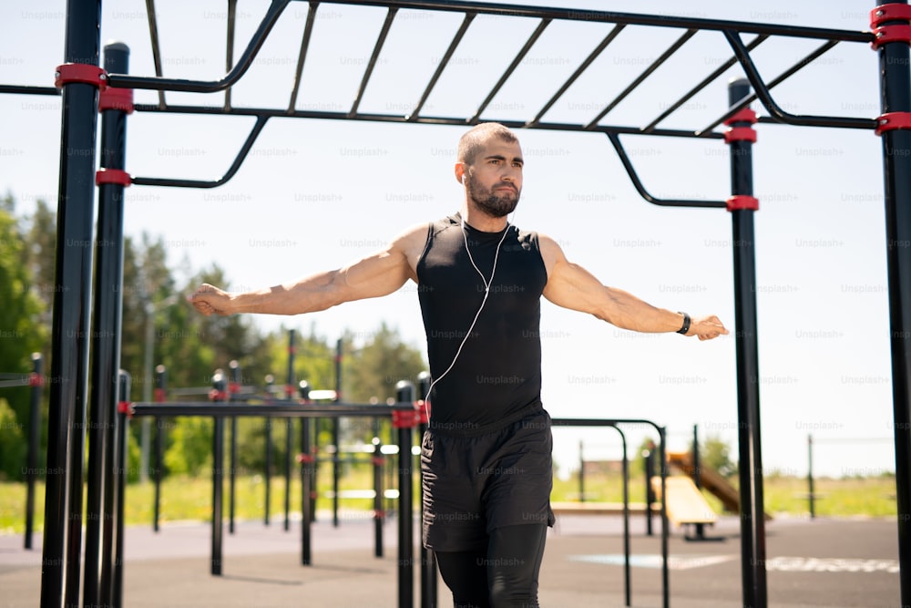 Strong young male athlete in black activewear outstretching his arms while exercising on outdoor sportsground