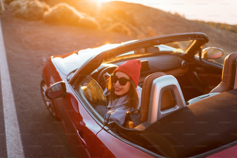 Mulher nova que viaja de carro em uma bela estrada da montanha durante um pôr do sol