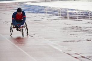Carreras en silla de ruedas. Atleta masculino discapacitado de voluntad fuerte en el entrenamiento de silla de ruedas deportiva en el estadio de atletismo al aire libre en un día lluvioso frío