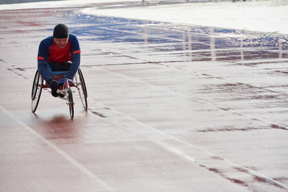 Wheelchair racing. Strong-willed handicapped male athlete in sport wheelchair training at outdoor track and field stadium on cold rainy day