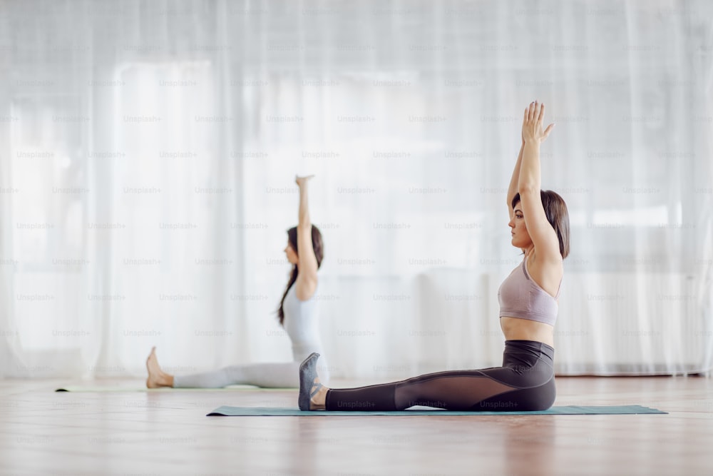Two beautiful fit girls sitting on the mat in yoga studio and doing yoga exercises. Hands are above the heads. Selective focus on the girl in foreground.