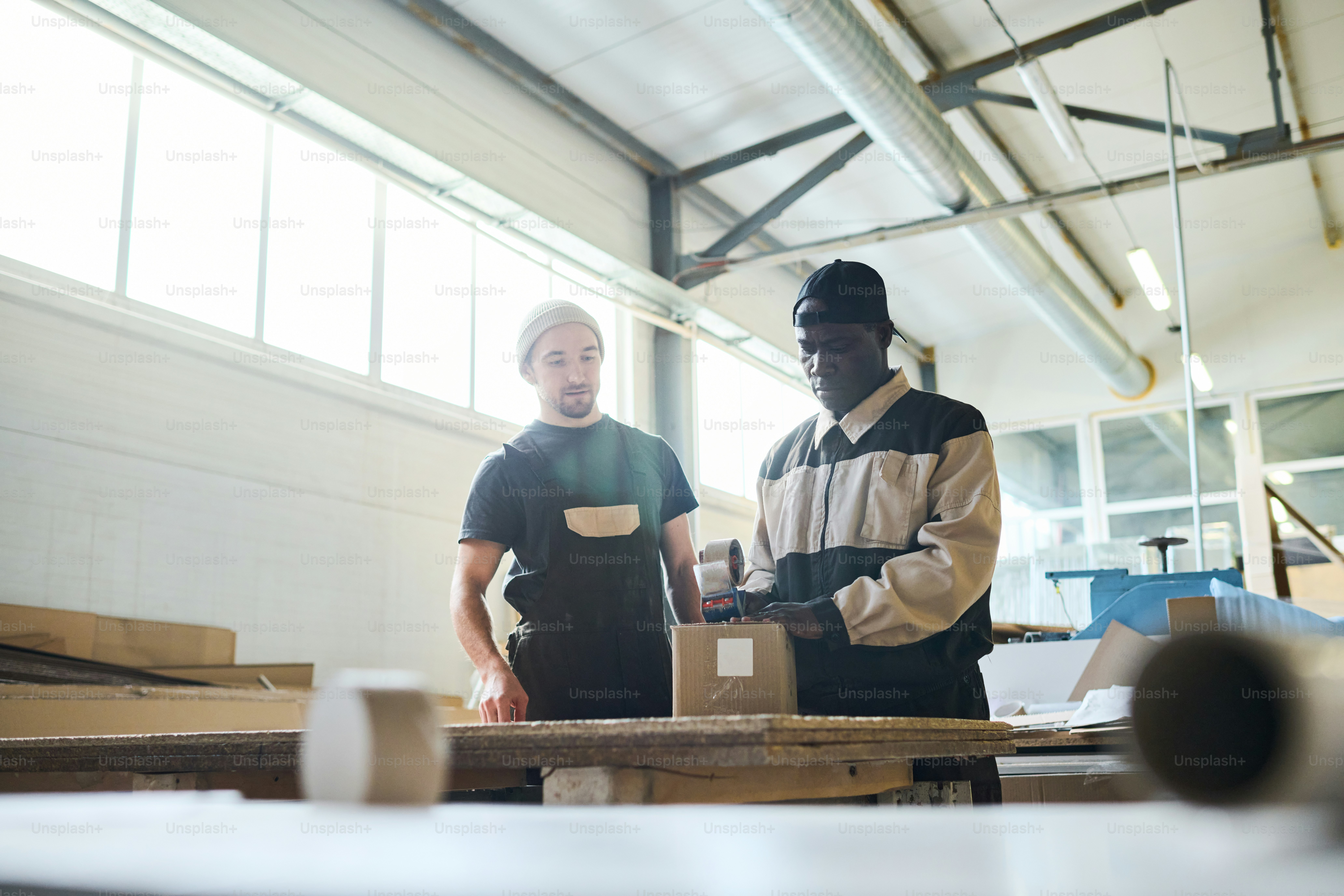 African worker in uniform packing box with products at table with assistant during their work at warehouse