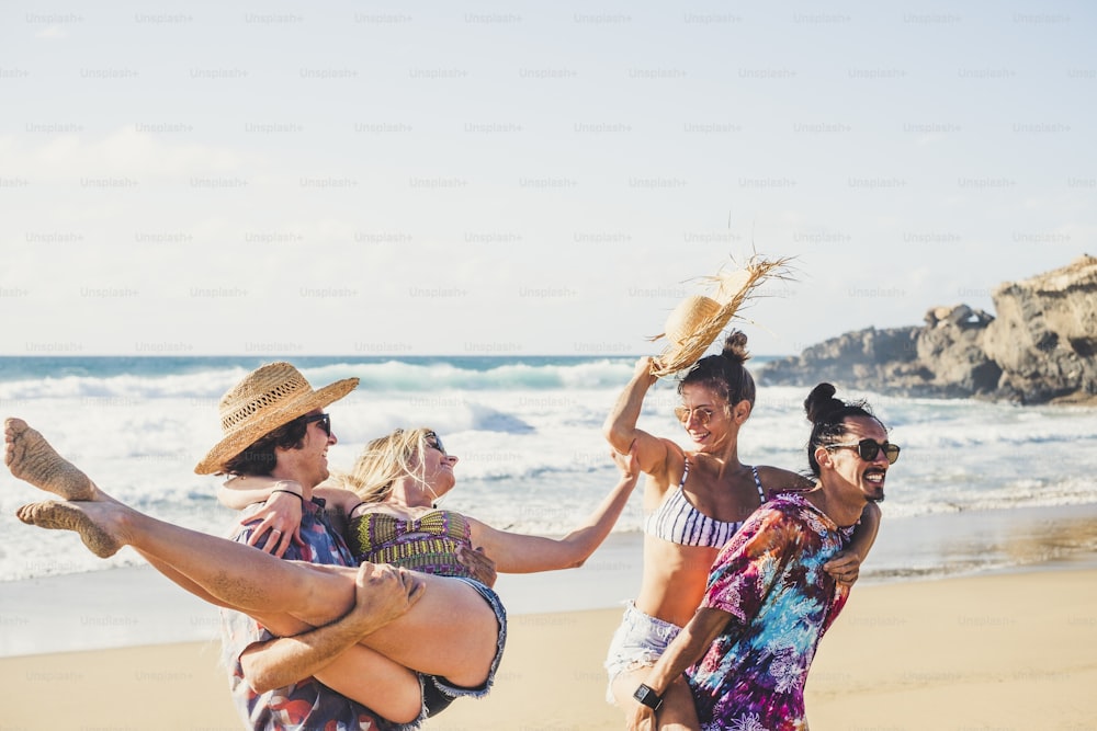 Cheerful group of people boys and girls have fun together at the beach during summer holiday vacation - men carrying women - coloured clotehs for outdoor sea lifestyle concept