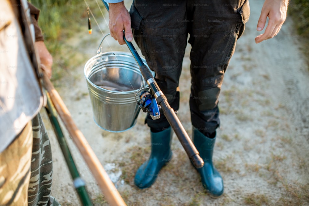 Men holding fishing rods and bucket with fresh caught fish outdoors. Close-up view with no face
