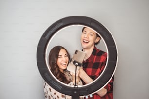 Young couple of beauty bloggers, hair stylist, hairdresser and model with hair style shoot on mobile phone with ring lamp on grey wall background