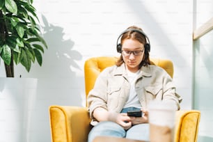 Young brunette teenager girl college or high school student in glasses using mobile phone, listen music in headphones in yellow chair at public place