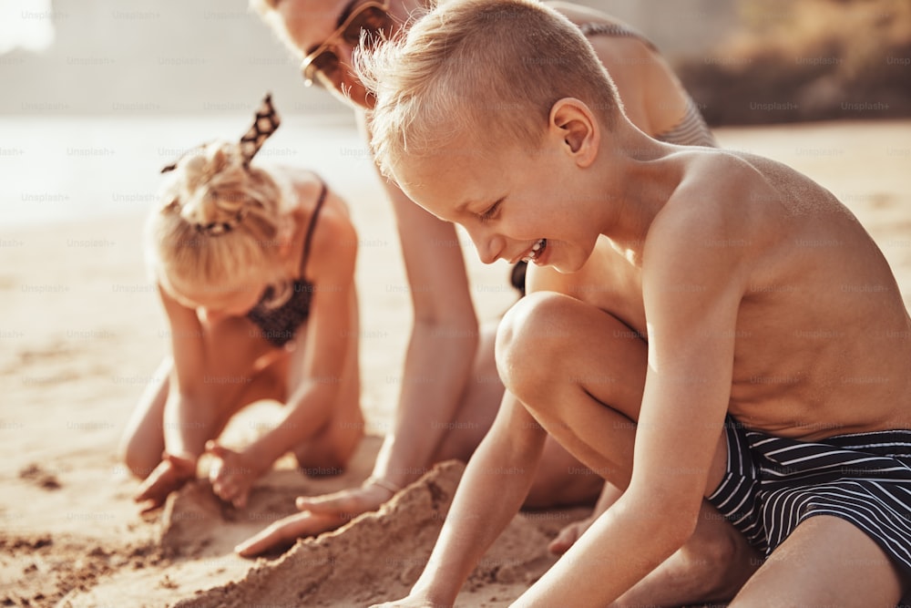 Ragazzino sveglio in costume da bagno che gioca nella sabbia con sua madre e sua sorella sullo sfondo durante una vacanza estiva in spiaggia