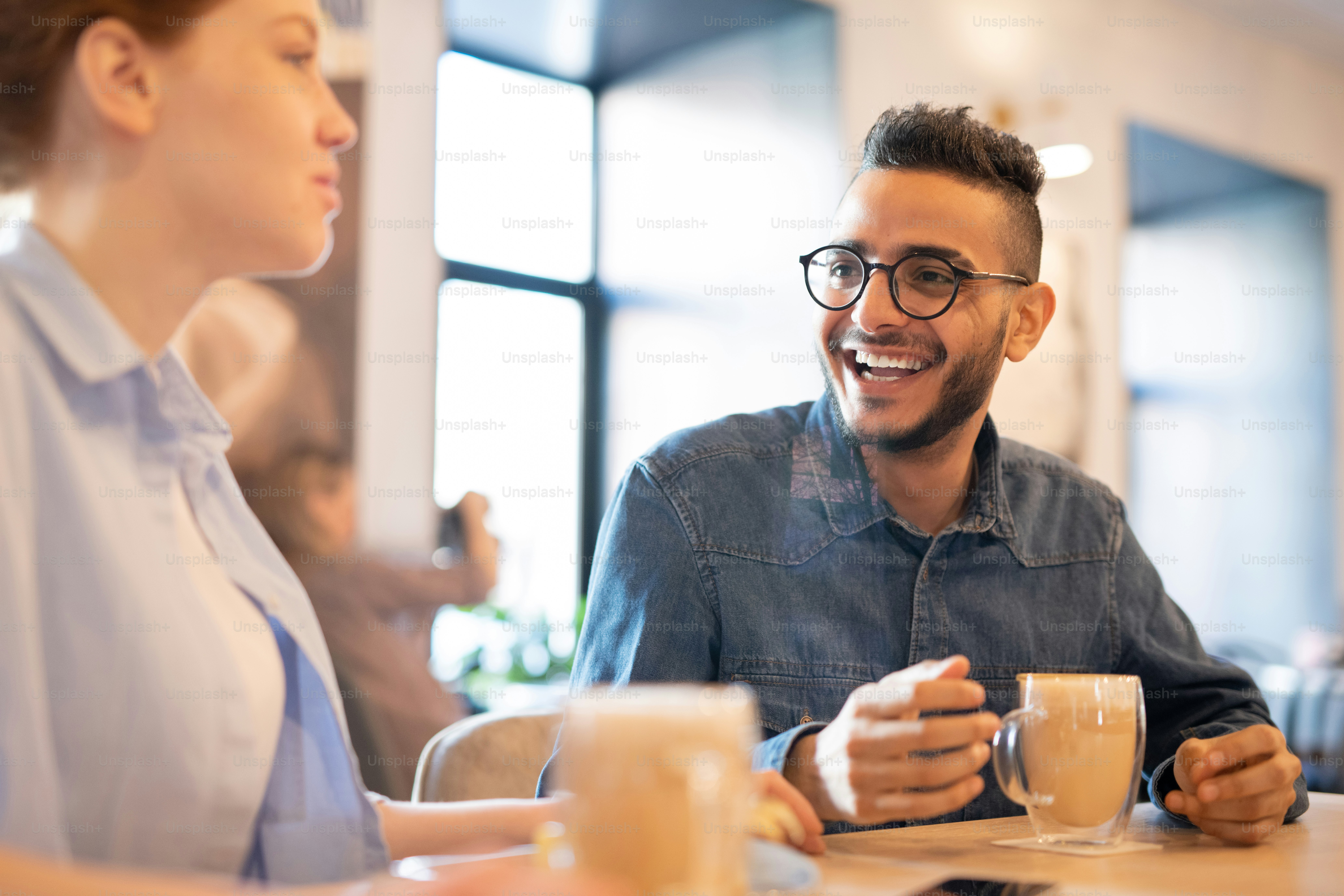 Young casual laughing man talking to his girlfriend while both having coffee in cozy cafe after college