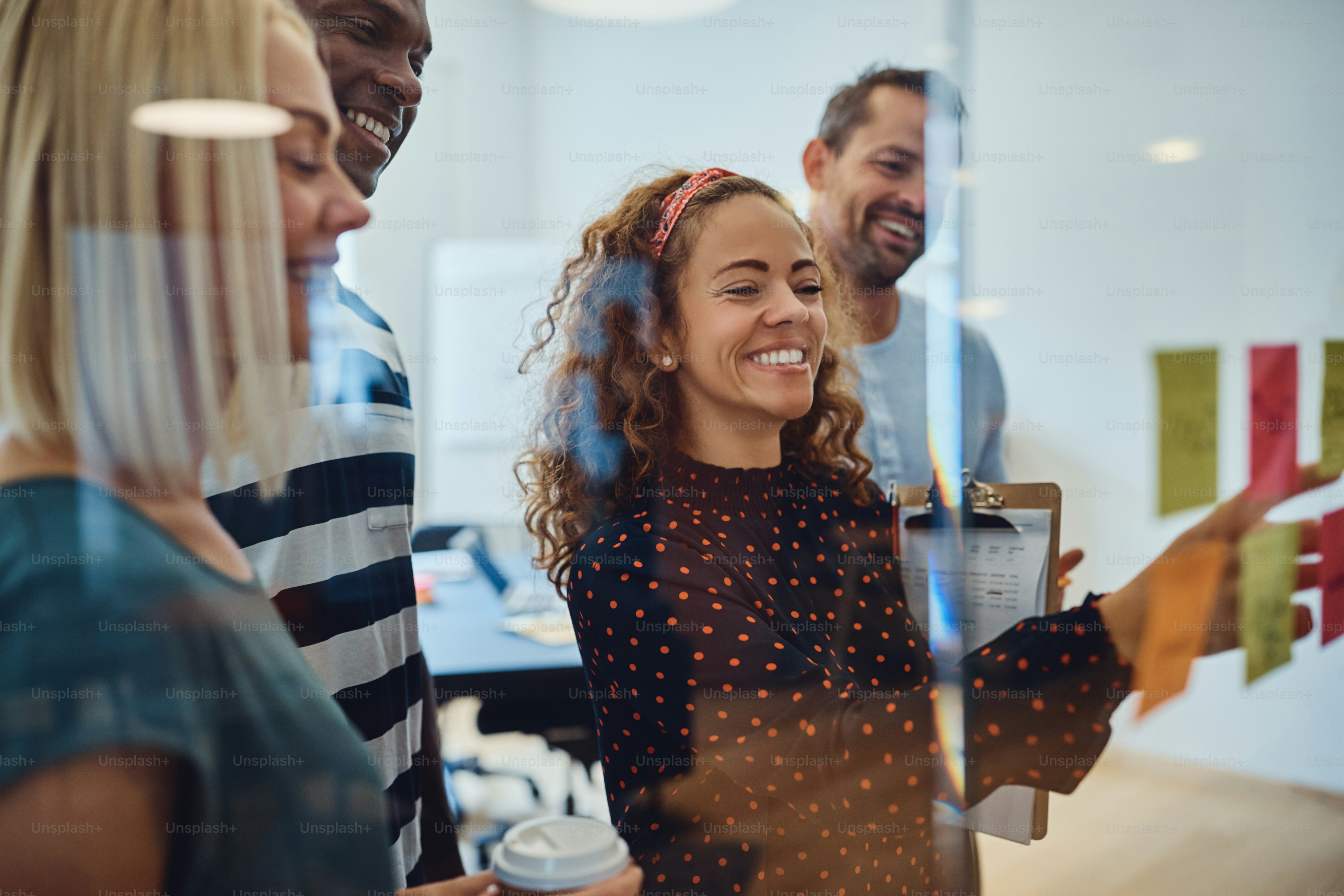 Diverse group of smiling young designers standing together in an office brainstorming with sticky notes on a glass wall