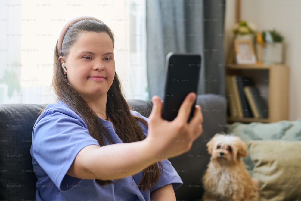 Mädchen mit Down-Syndrom hält Smartphone vor sich, während es Selfie oder Videoanruf gegen flauschiges Haustier im Wohnzimmer macht