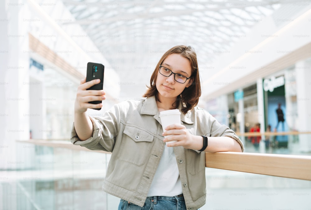 Giovane studentessa adolescente bruna in occhiali usando il telefono cellulare che scatta selfie con tazza di caffè di carta al centro commerciale del luogo pubblico