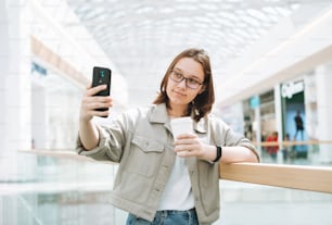 Young brunette teenager girl student in glasses using mobile phone taking selfie with paper coffee cup at public place shopping mall