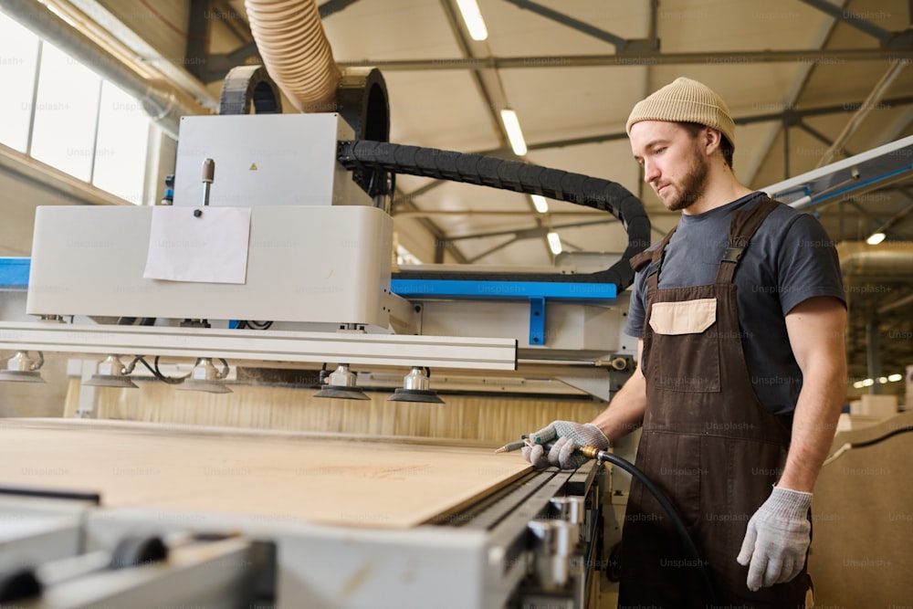 Jeune charpentier en tablier travaillant avec des planches de bois à la machine moderne à l’usine de meubles