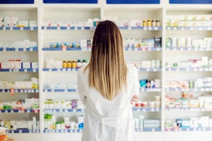 Female apothecary worker with backs turned standing at pharmacy and looking at medicines and drugs.