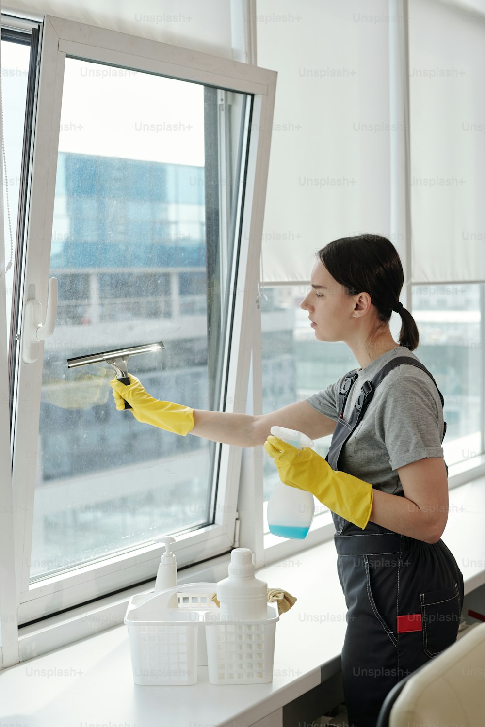 젊은 여성 클리너는 창문에 세제를 뿌리고 대형 오픈 스페이스 사무실에서 일하는 동안 특수 실리콘 주걱으로 문지릅니다.