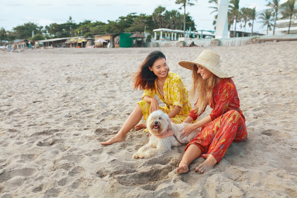 M�ädchen mit Hund am Strand. Models in böhmischer Kleidung und Strohhut mit Haustier an der sandigen Küste. Schöne Frauen in Maxikleidern genießen es, sich am Meeresufer auszuruhen. Boho-Stil für modischen Look im Resort.