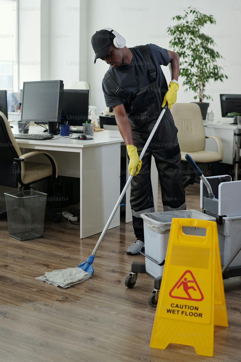 Young African American male janitor in headphones bending forwards while washing floor in large contemporary openspace office
