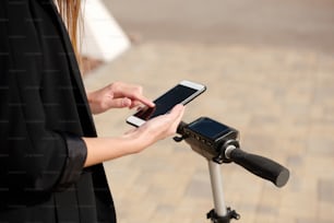 Hands of young female scrolling in smartphone while standing on electric scooter and listening to favorite music on her way to work