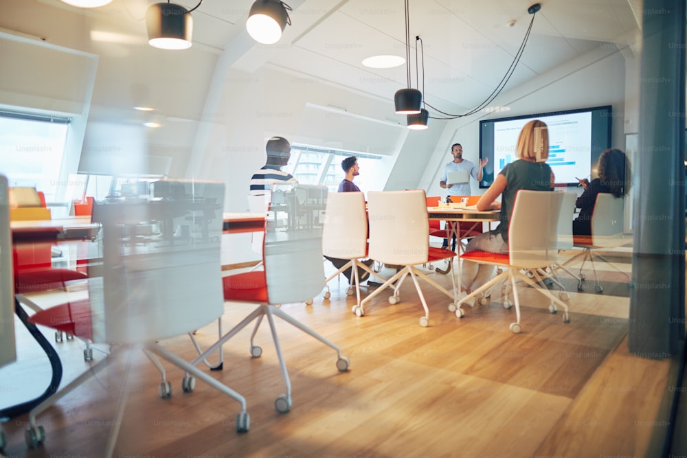 Manager discutant de graphiques sur un écran lors d’une réunion avec un groupe diversifié de collègues assis autour d’une table à l’intérieur d’un bureau en verre