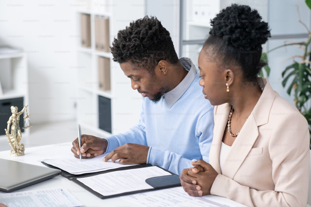 Serious young Blackman sitting at table in lawyers office and signing divorce papers