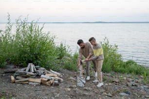 Giovane uomo che mostra a suo figlio adolescente come tagliare la legna da ardere in riva all'acqua