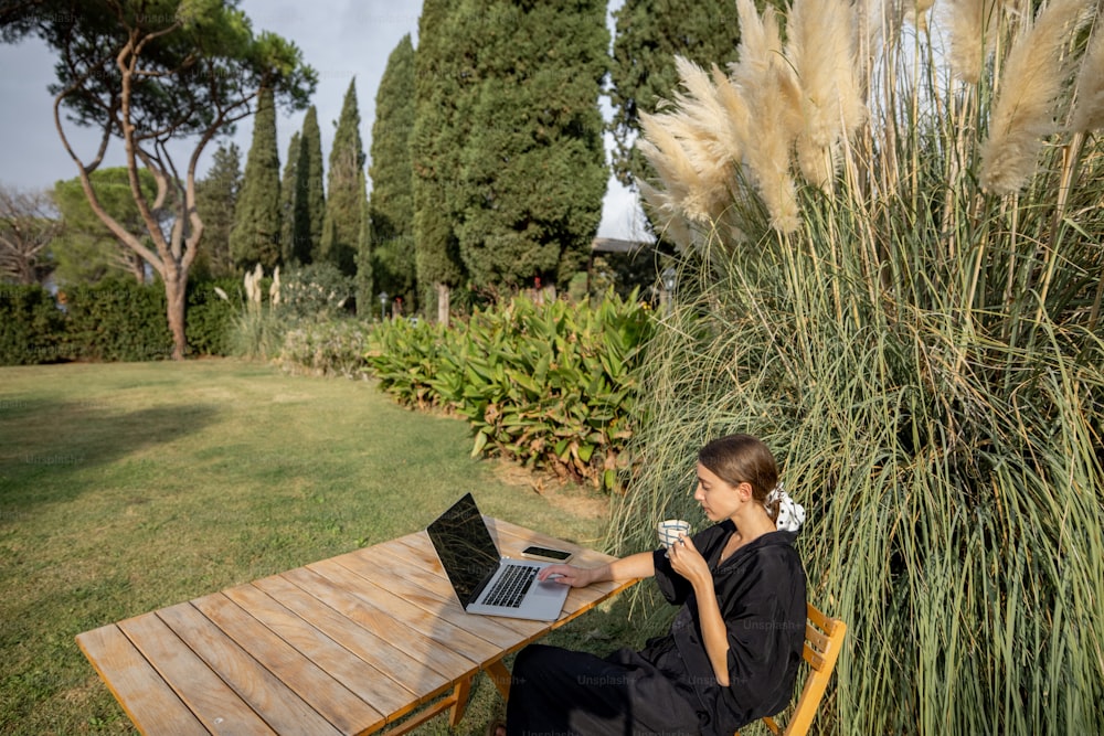 Joven mujer caucásica mirando algo en una computadora portátil y bebiendo té mientras está sentada en una mesa de madera en el césped verde mediterráneo. Concepto de fin de semana, descanso y vacaciones. Día soleado