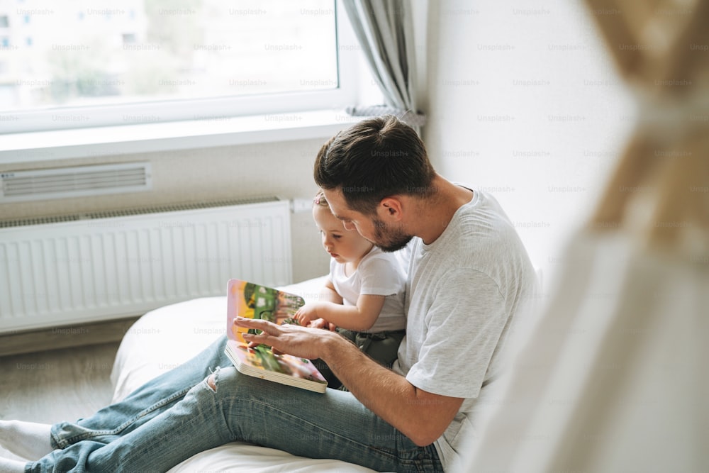 Happy father young man and baby girl little daughter having fun reading a book in children room at home